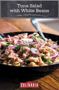 Tuna salad with white beans, red onion, and parsley in a black bowl with a fork and spoon, on a wooden table.