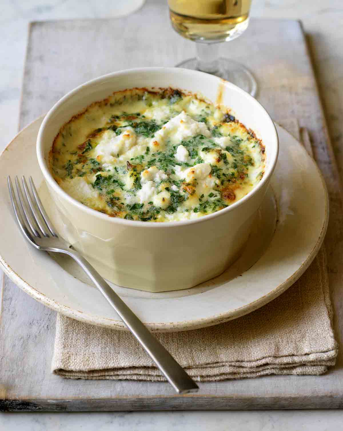 En vit gratängform fylld med en krämig zucchinigratäng, toppad med getost, på en tallrik med en gaffel vid sidan av.