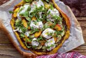 A whole zucchini, peach, and burrata pizza, topped with arugula, on a piece of parchment on a wooden surface.