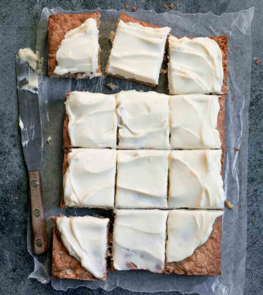 A whole apple cake, covered in cream cheese frosting and cut into 12 squares on a sheet of wax paper with a knife on the side.