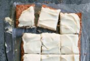 A whole apple cake, covered in cream cheese frosting and cut into 12 squares on a sheet of wax paper with a knife on the side.