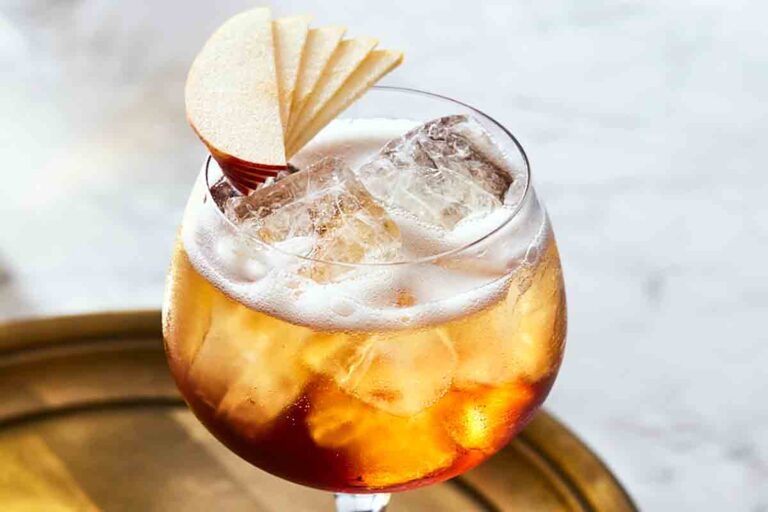 A wine glass filled with ice, amber liquid, a little foam on top, and an apple fan garnish.