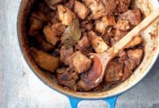 A blue Dutch oven filled with chunks of braised pork and a bay leaf with a wooden spoon resting inside.