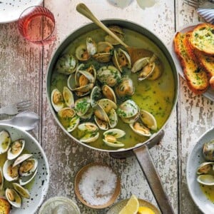 A pot filled with opened clams in garlic butter with bowls of clams, lemon wedges, and a plater of toasted sourdough beside it.