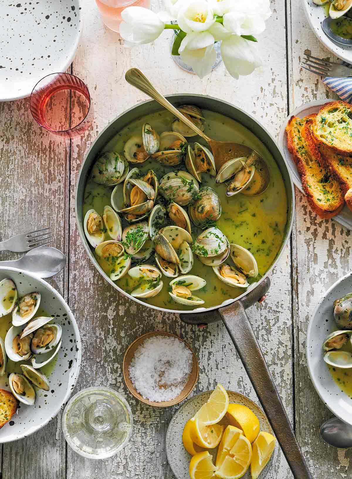 A pot filled with opened clams in garlic butter with bowls of clams, lemon wedges, and a plater of toasted sourdough beside it.