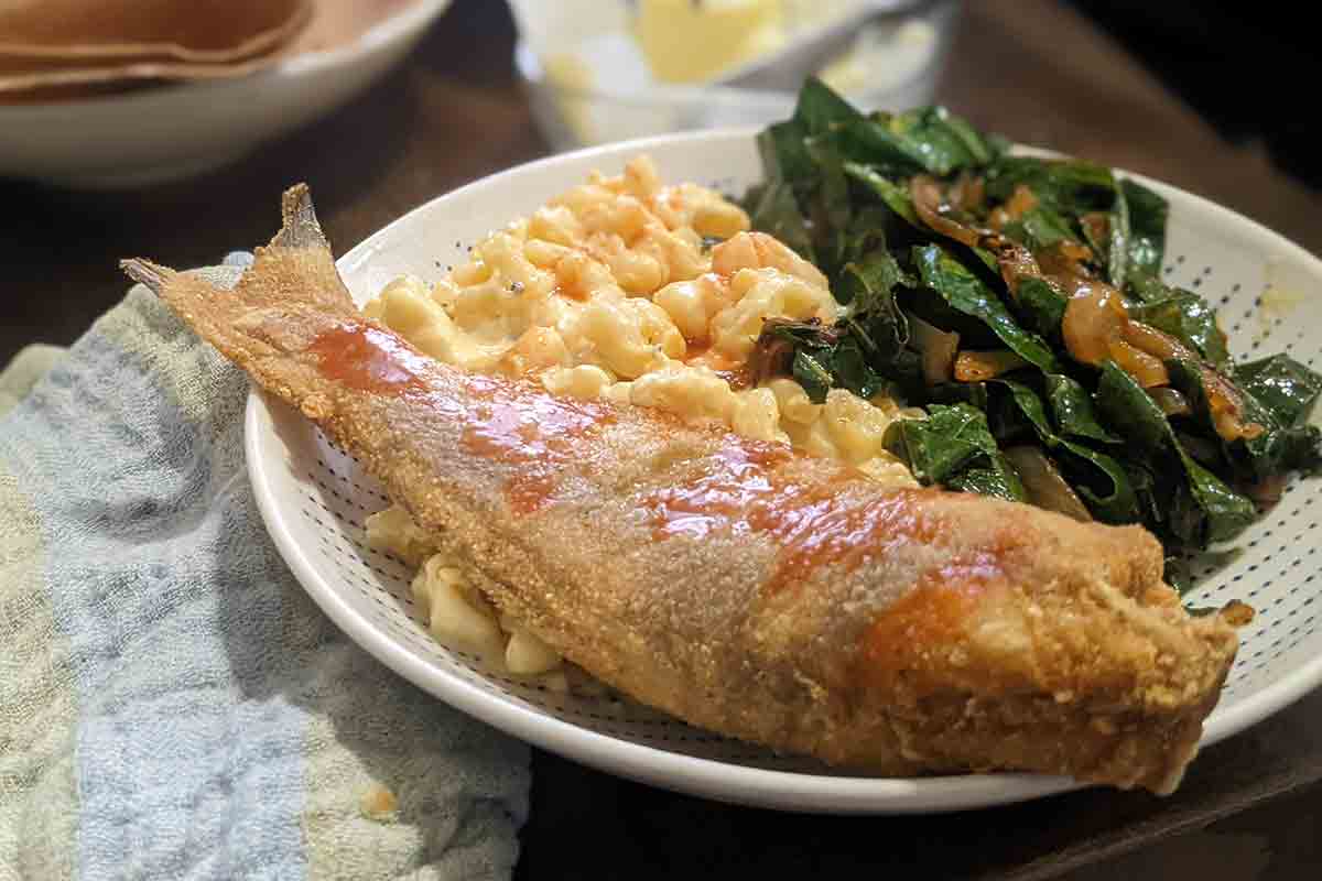 A serving of mac and cheese on a plate with fried fish and collard greens.