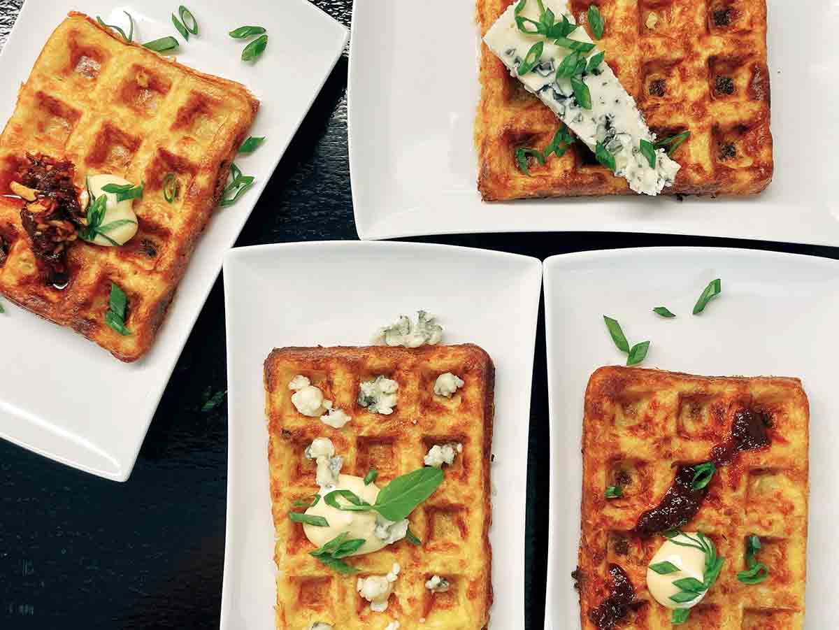 Four hash brown waffles on white rectangular plates topped with a variety of cheeses, aiolis, and tapenades.