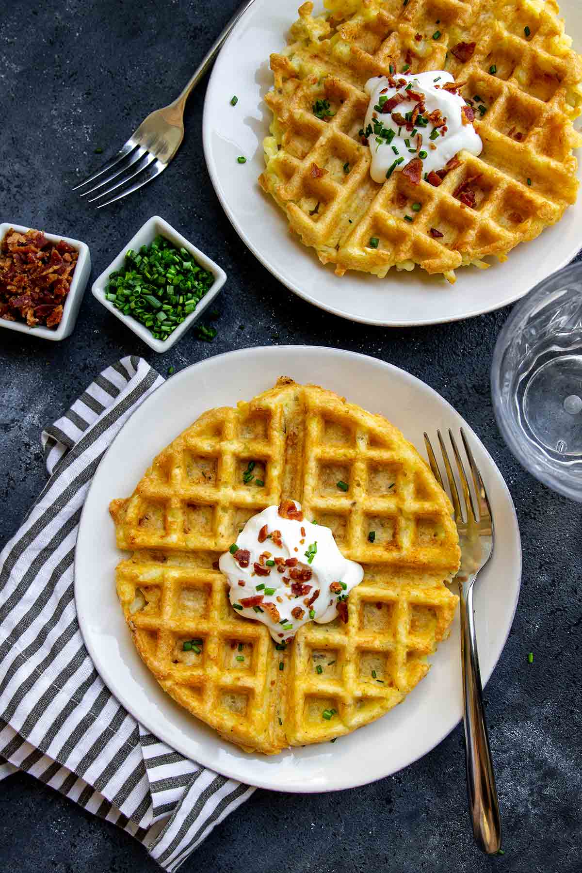 Two round hash brown waffles on white plates, topped with sour cream, bacon, and chives.