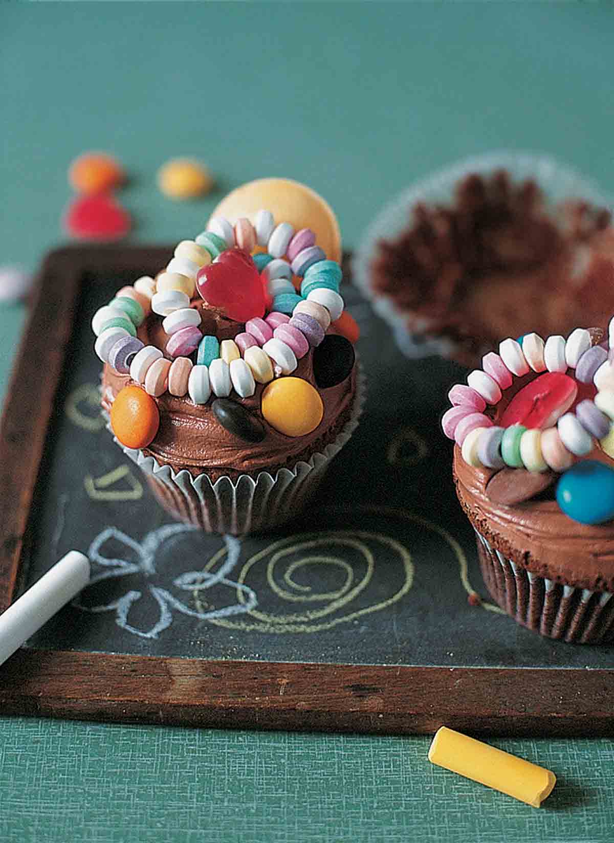 Chocolate cupcakes topped with chocolate frosting and pieces of candy on a children's chalkboard with pieces of chalk on the side.
