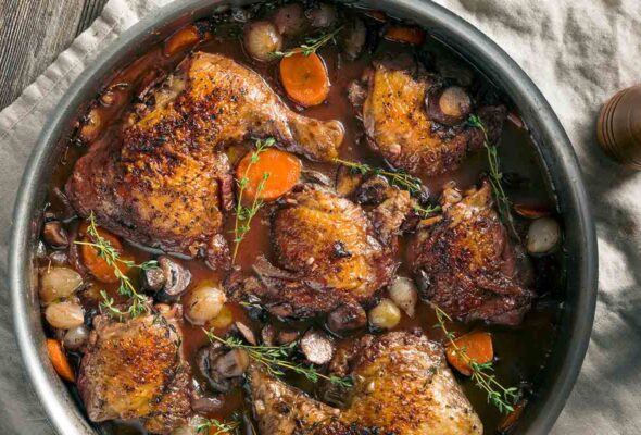 A metal pot filled with braised chicken, pearl onions, mushrooms, carrots, and thyme sprigs.