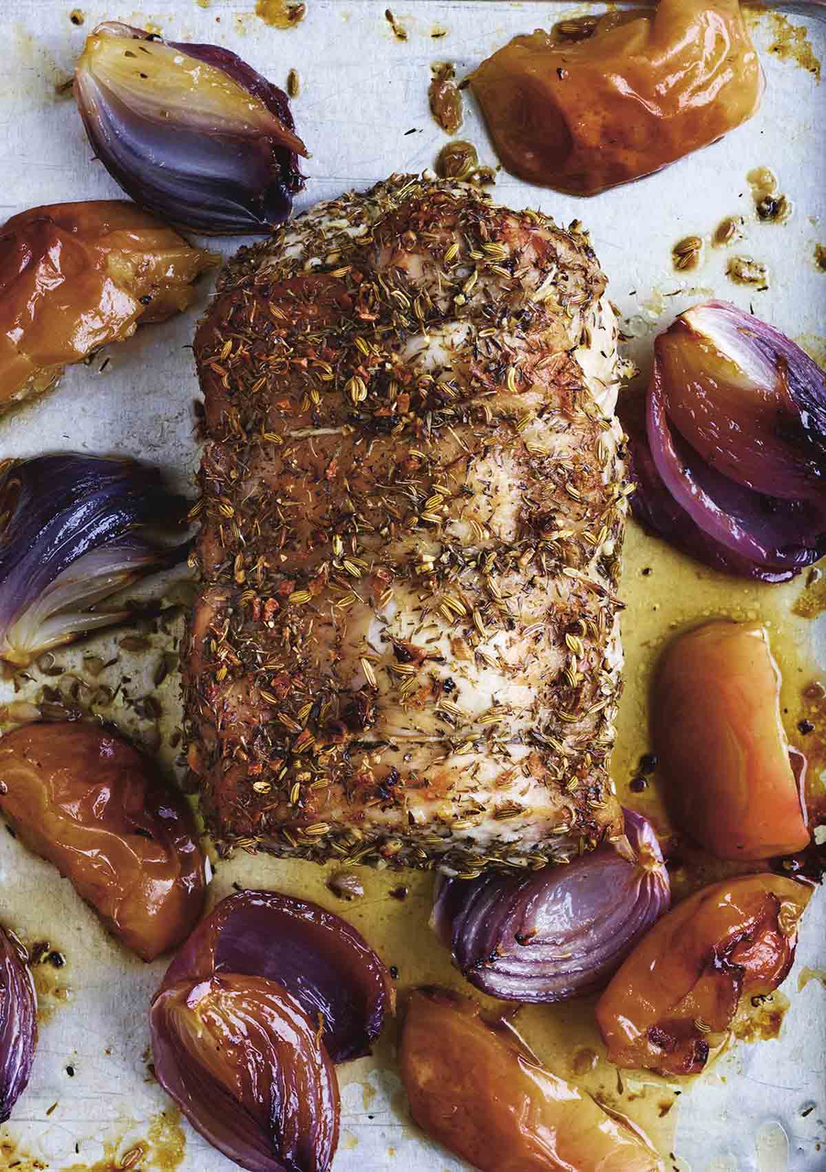 A roast pork loin with apples and onions scattered around it on a baking sheet.