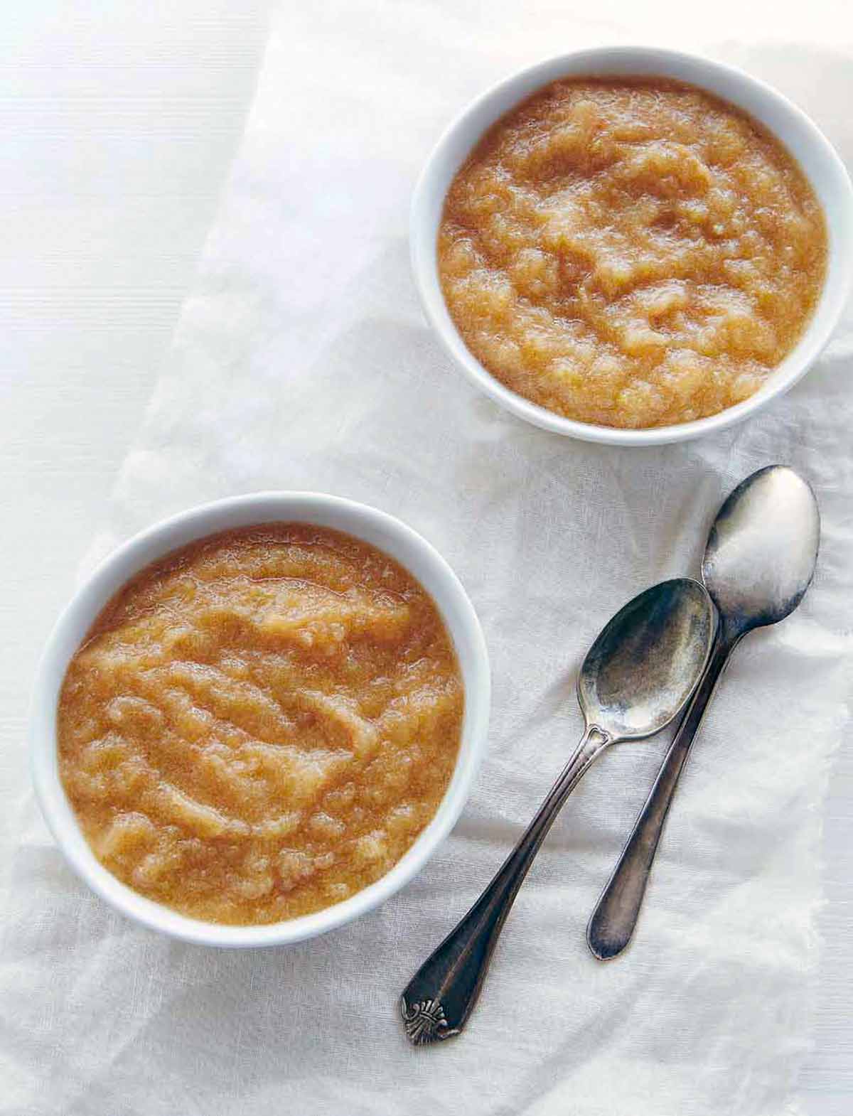 Two bowls of slow cooker applesauce with two spoons nearby.