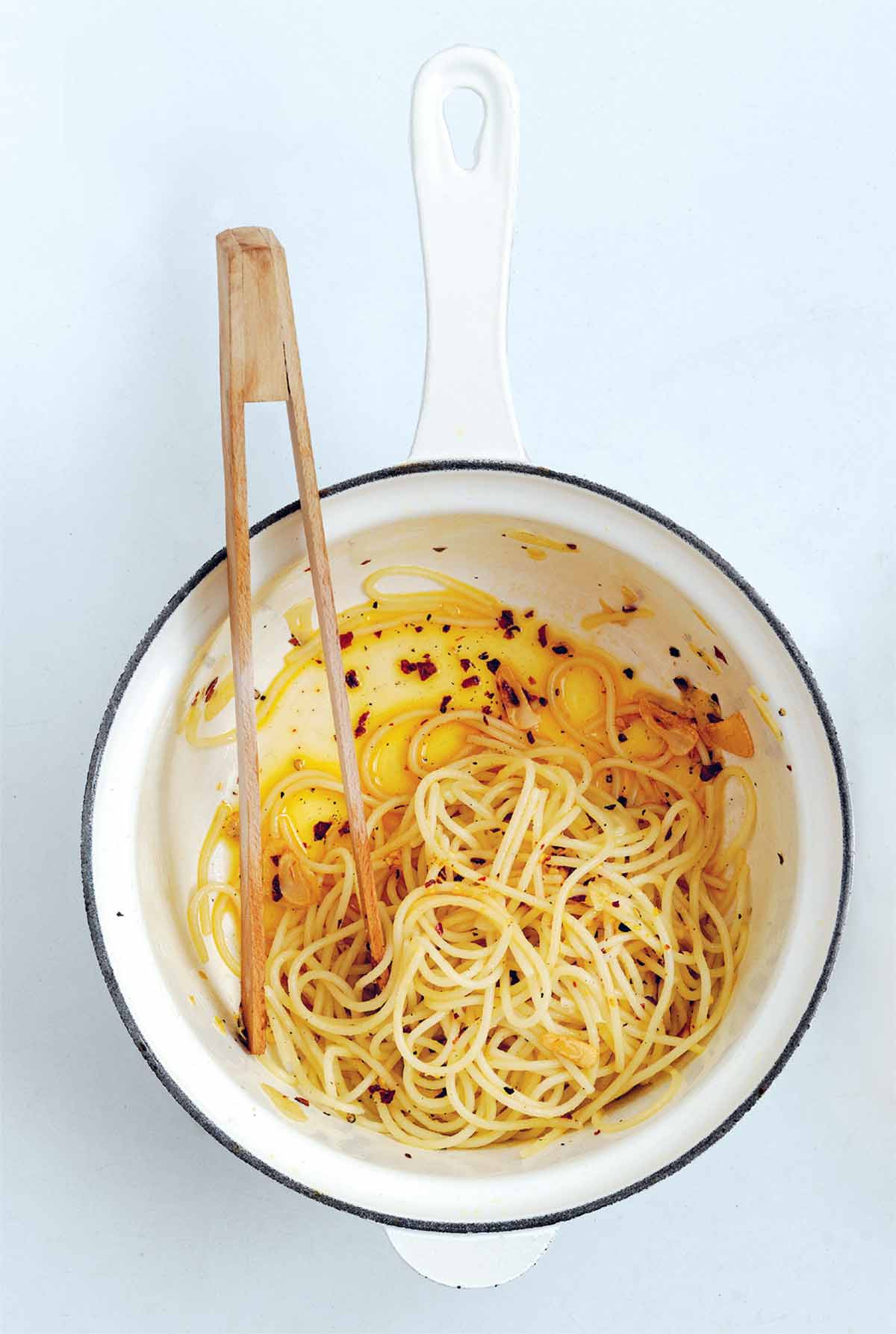 En vit emaljkruka med blå kant med spagetti, chiliflakes och olja i botten.  En uppsättning trätång sitter inuti.