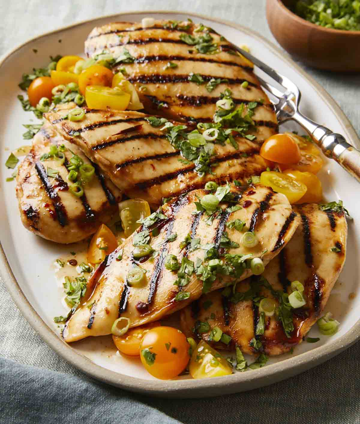 Five chicken breast pieces with grill marks on a platter, topped with halved cherry tomatoes, scallions, and cilantro.