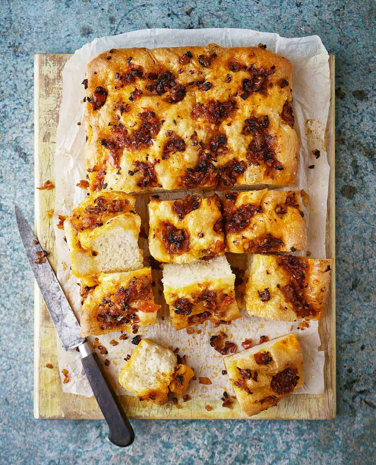 A partially cut loaf of tomato focaccia on a sheet of parchment on a wooden cutting board with a knife on the side.