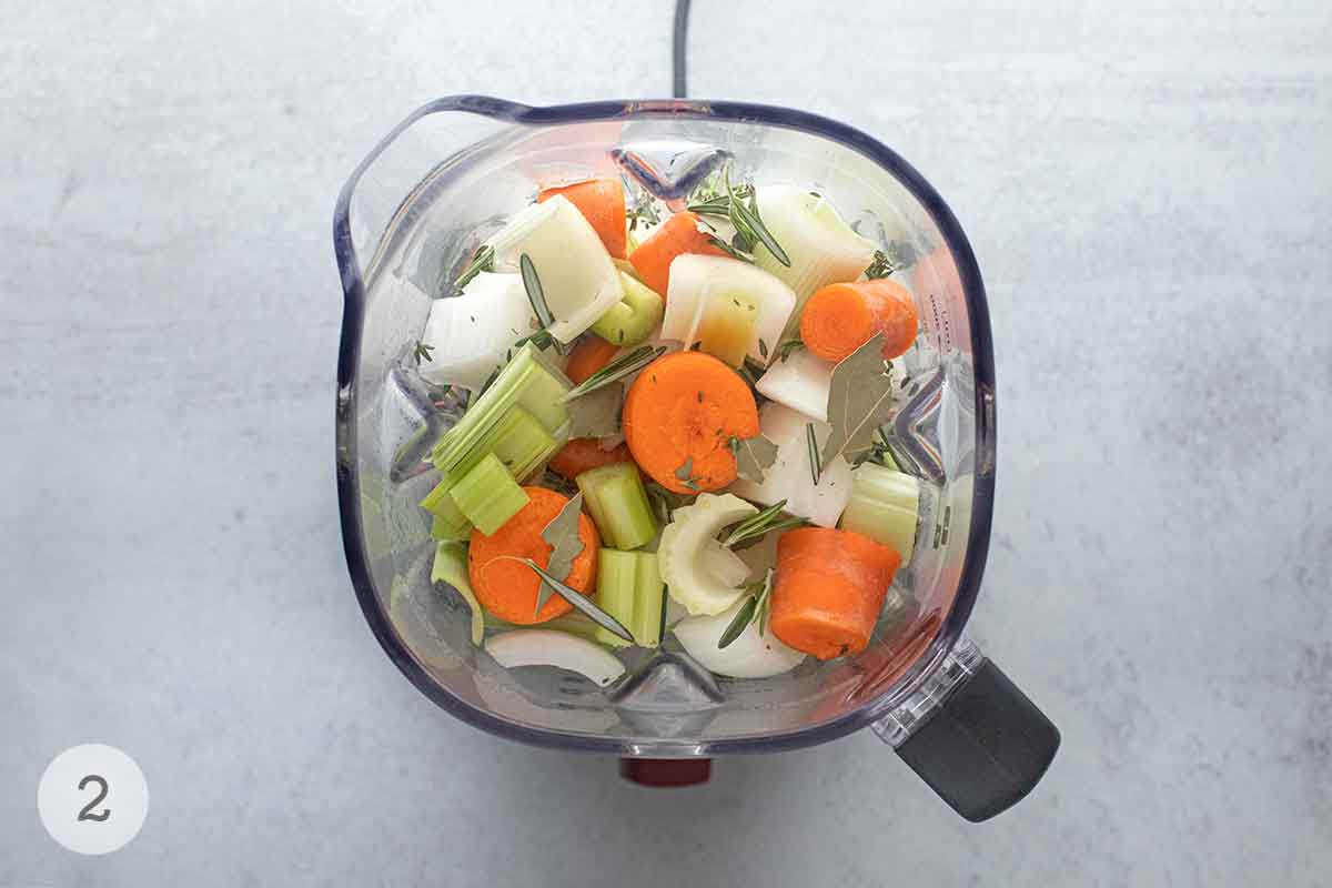 A blender filled with chopped carrots, onion, celery, leeks, rosemary, and thyme.