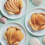 Three fanned pear tarts on puff pastry on white plates with seashells scattered around.