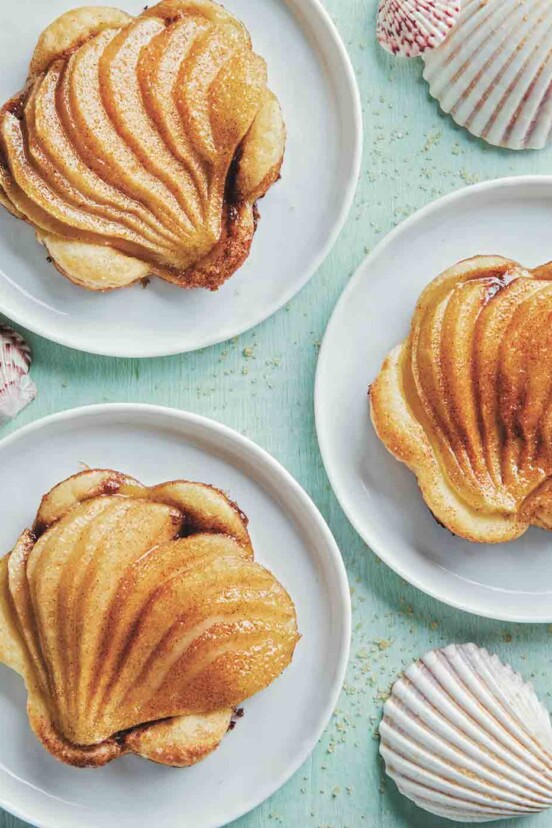 Three fanned pear tarts on puff pastry on white plates with seashells scattered around.