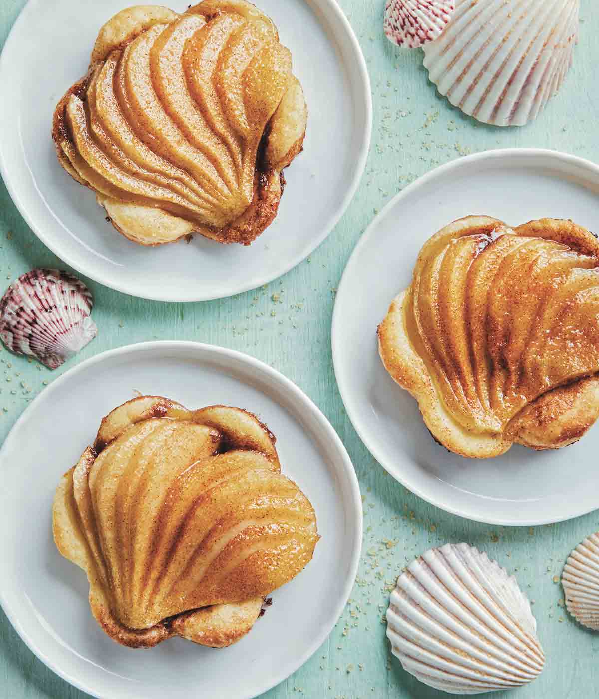 Three fanned pear tarts on puff pastry on white plates with seashells scattered around.