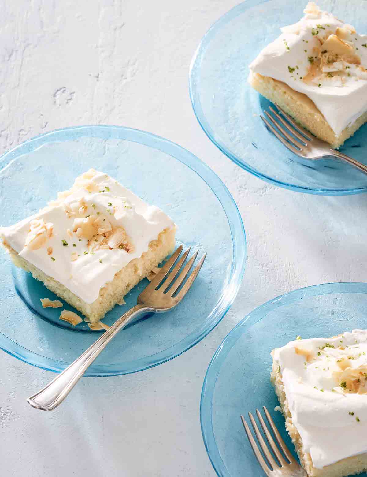 Three pieces of white cake topped with frosting and toasted coconut on individual blue glass plates with forks resting on the side.