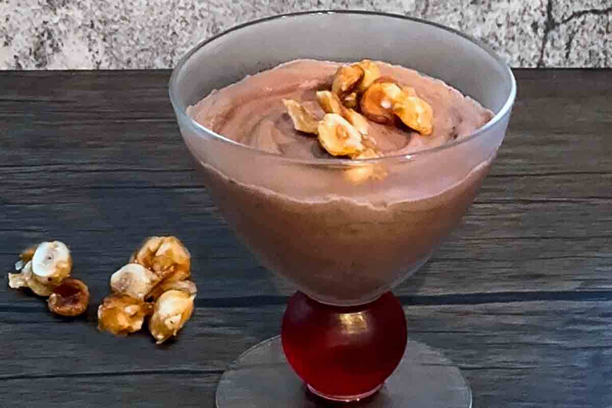 A dessert dish filled with dark chocolate mousse, topped with candied hazelnuts and more hazelnuts on the side.
