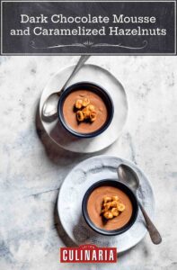 Två skålar fyllda med chokladmousse och kanderade hasselnötter ovanpå, sittande på tallrikar med en silversked på sidan av varje skål.