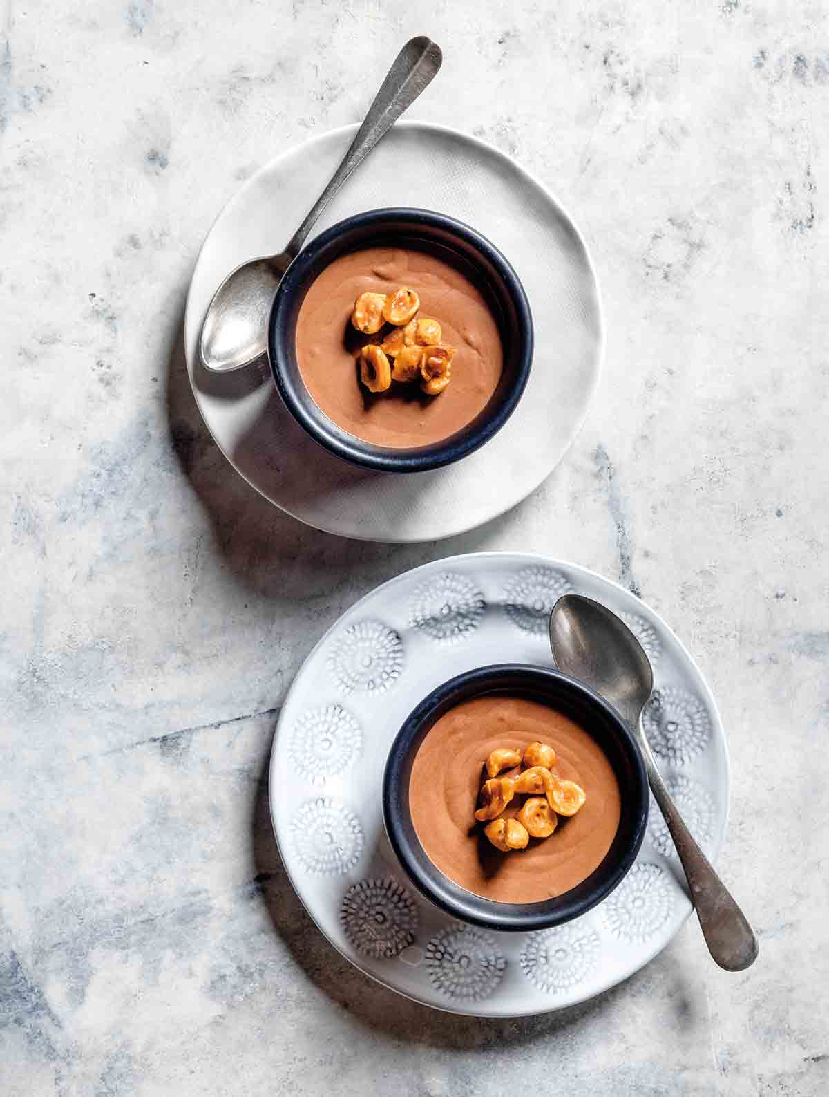 Two bowls filled with chocolate mousse and candied hazelnuts on top, sitting on plates with a silver spoon on the side of each bowl.