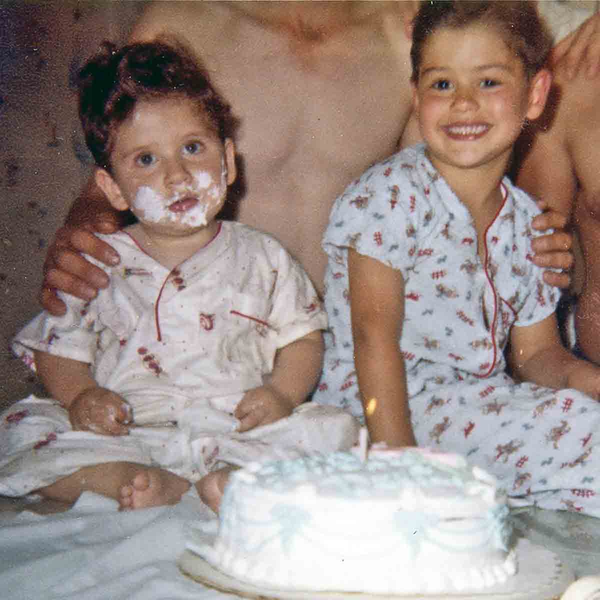 Two small boys in front of a birthday cake. One boy has frosting all over his face.