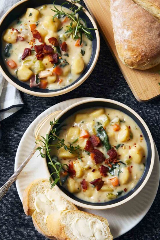 Two bowls of gnocchi soup, garnished with bacon and rosemary, with crusty bread on the side.