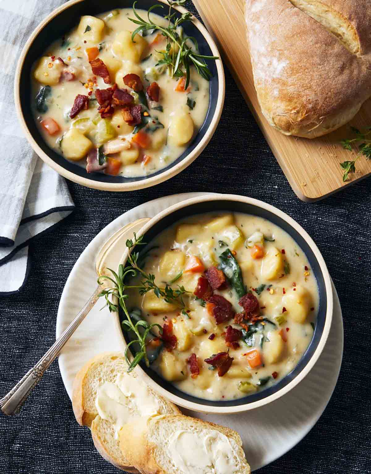 Two bowls of gnocchi soup, garnished with bacon and rosemary, with crusty bread on the side.