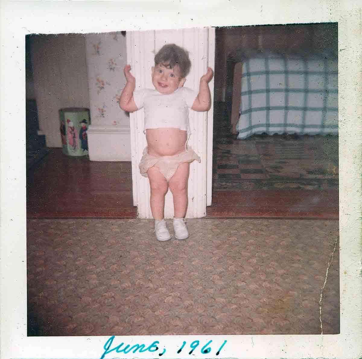 A small boy standing in his diaper between two bedroom doors.