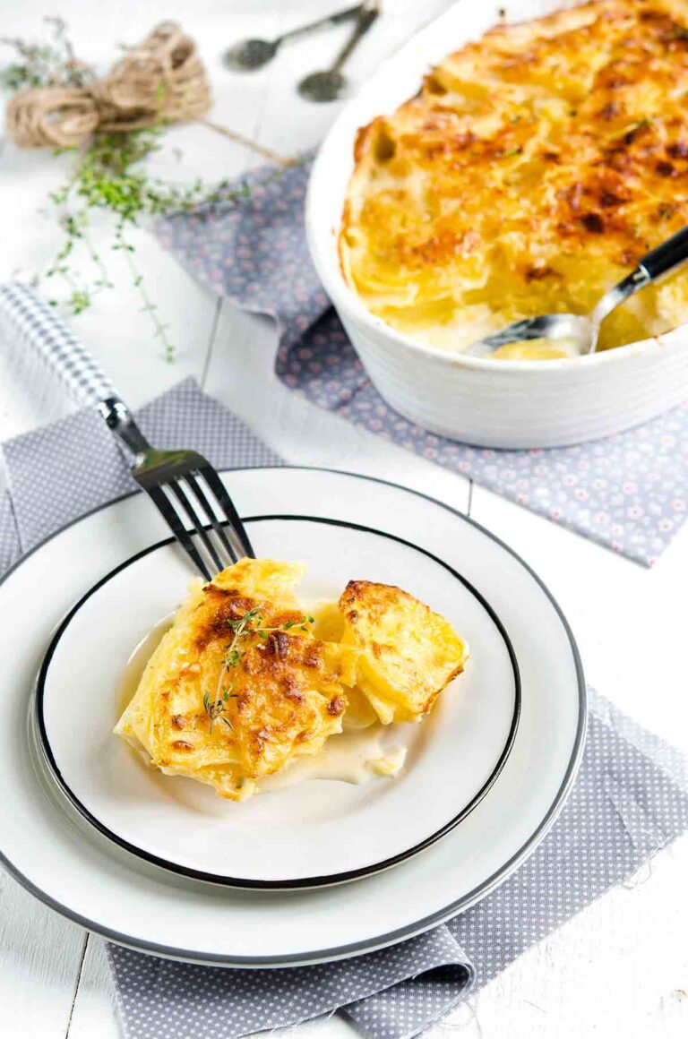 A round baking dish filled with scalloped potatoes and two stacked white plates topped with a serving of the scalloped potatoes.