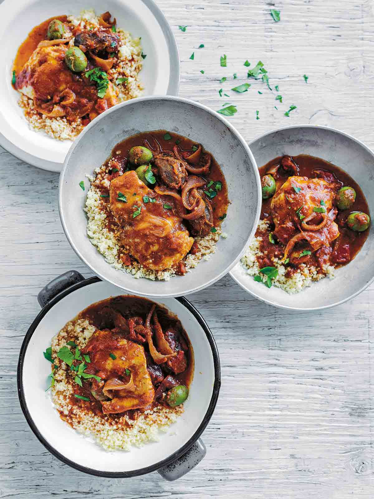 Four bowls of slow cooker chicken tagine, served over couscous with olives, and garnished with parsley.