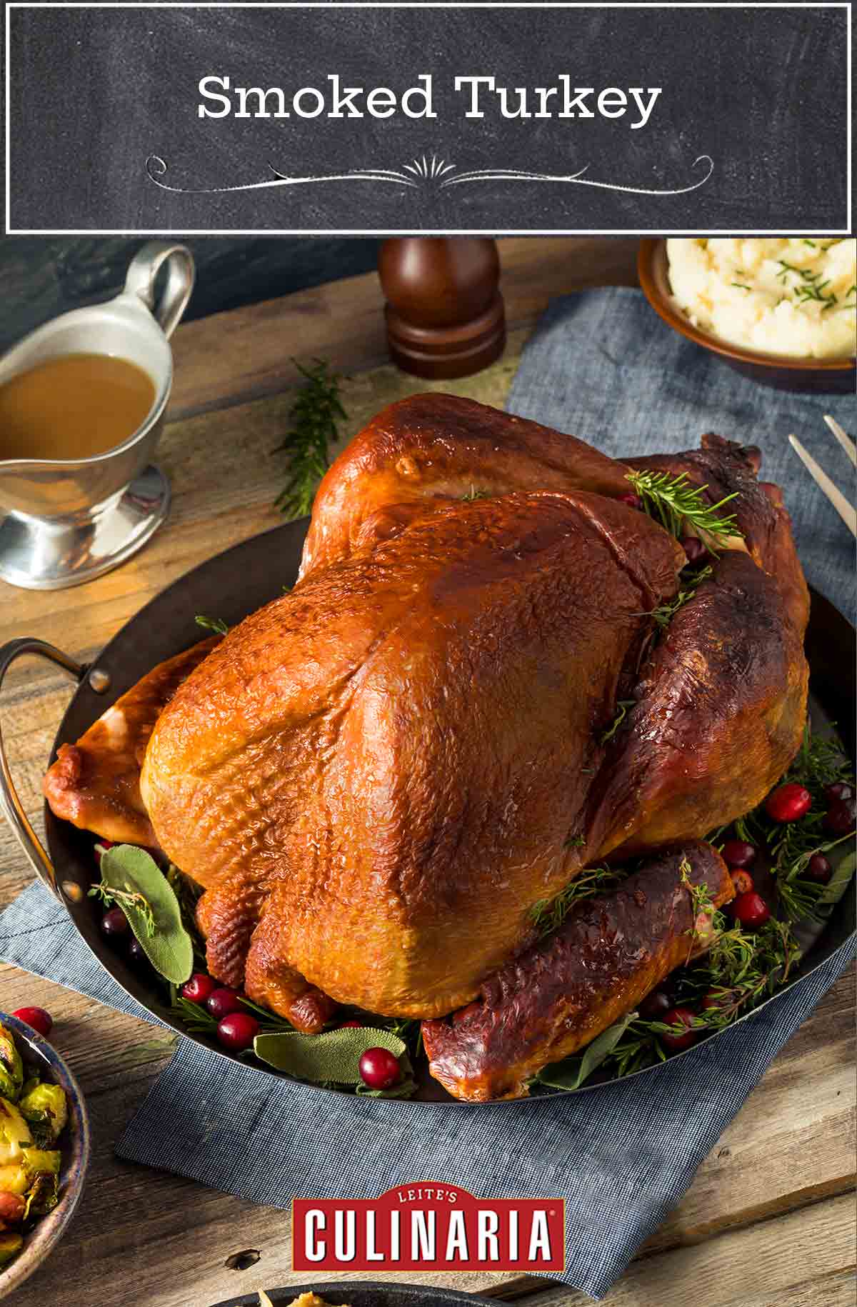 A whole smoked turkey in a metal serving dish on a bed of fresh herbs and cranberries.
