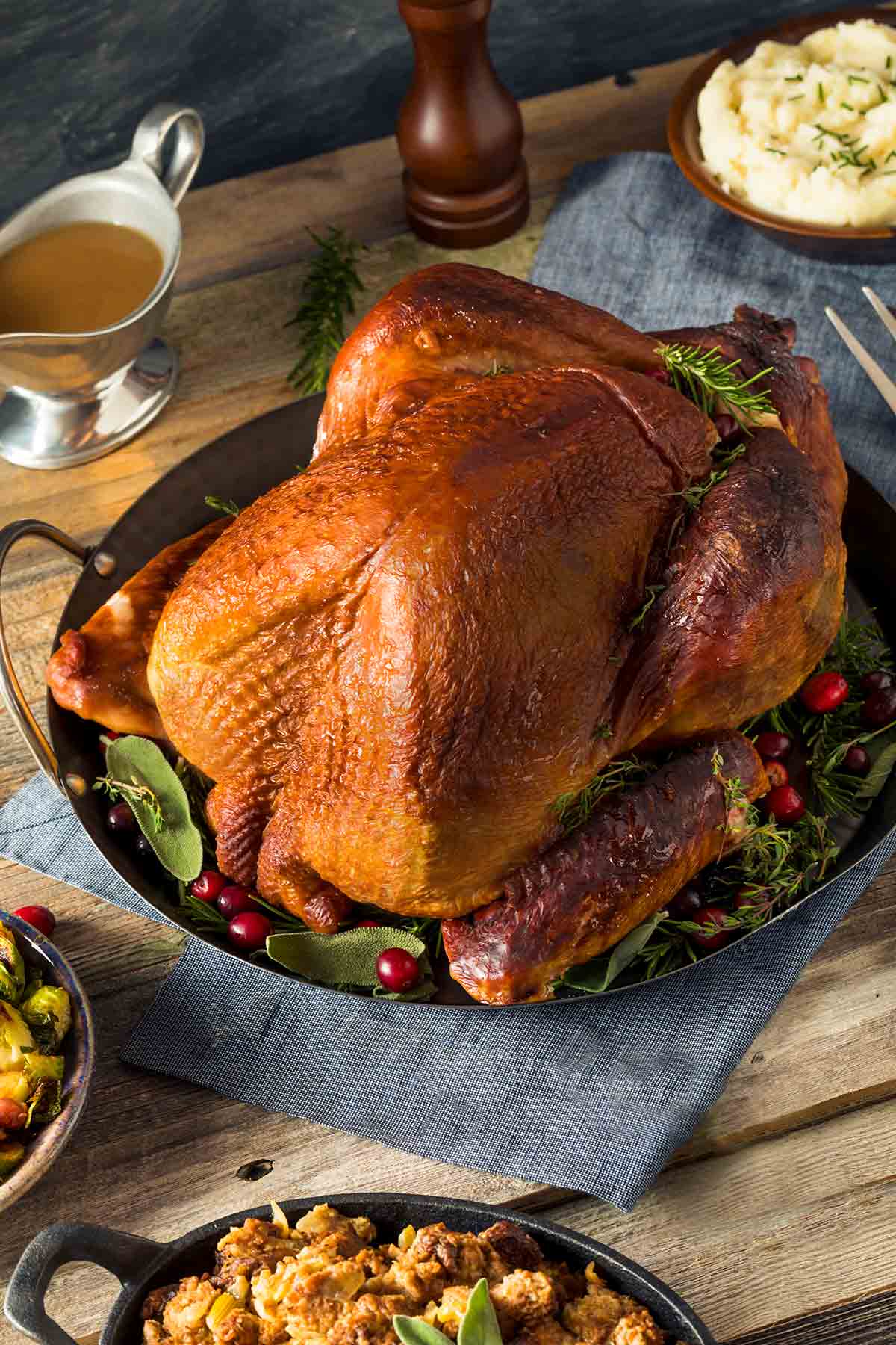 A whole smoked turkey in a metal serving dish on a bed of fresh herbs and cranberries.