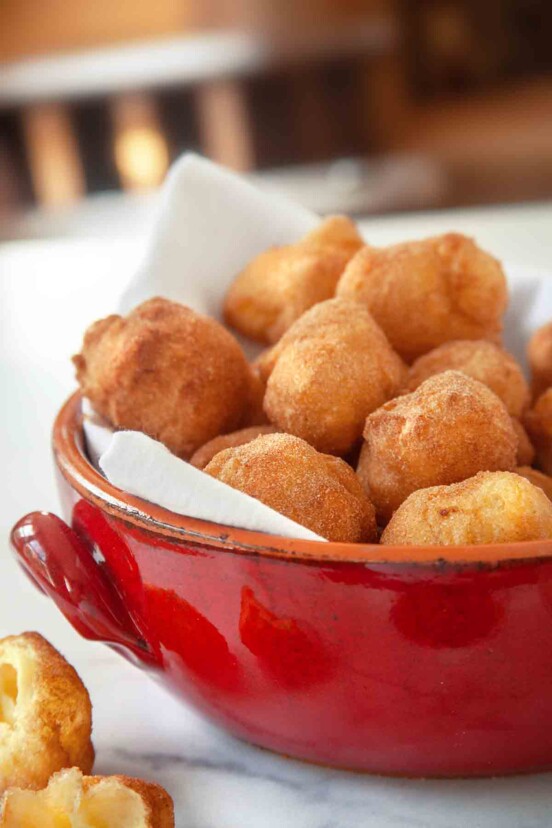 A red pot filled with sonhos dusted with cinnamon sugar and one cut Portuguese doughnut resting beside the bowl.