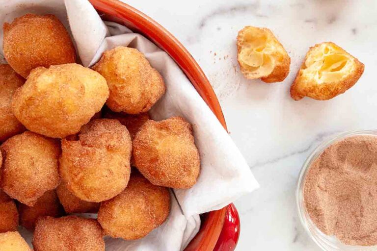 A red pot filled with sonhos dusted with cinnamon sugar and one cut Portuguese doughnut resting beside the bowl.