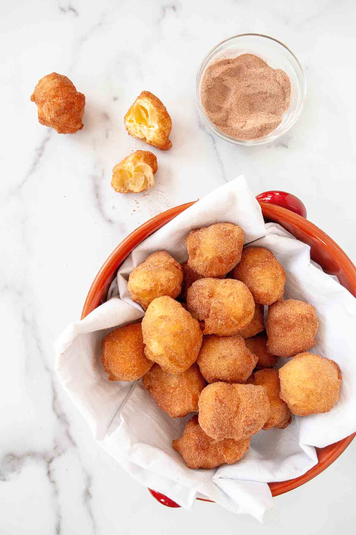 A red pot filled with sonhos dusted with cinnamon sugar and one cut Portuguese doughnut resting beside the bowl.