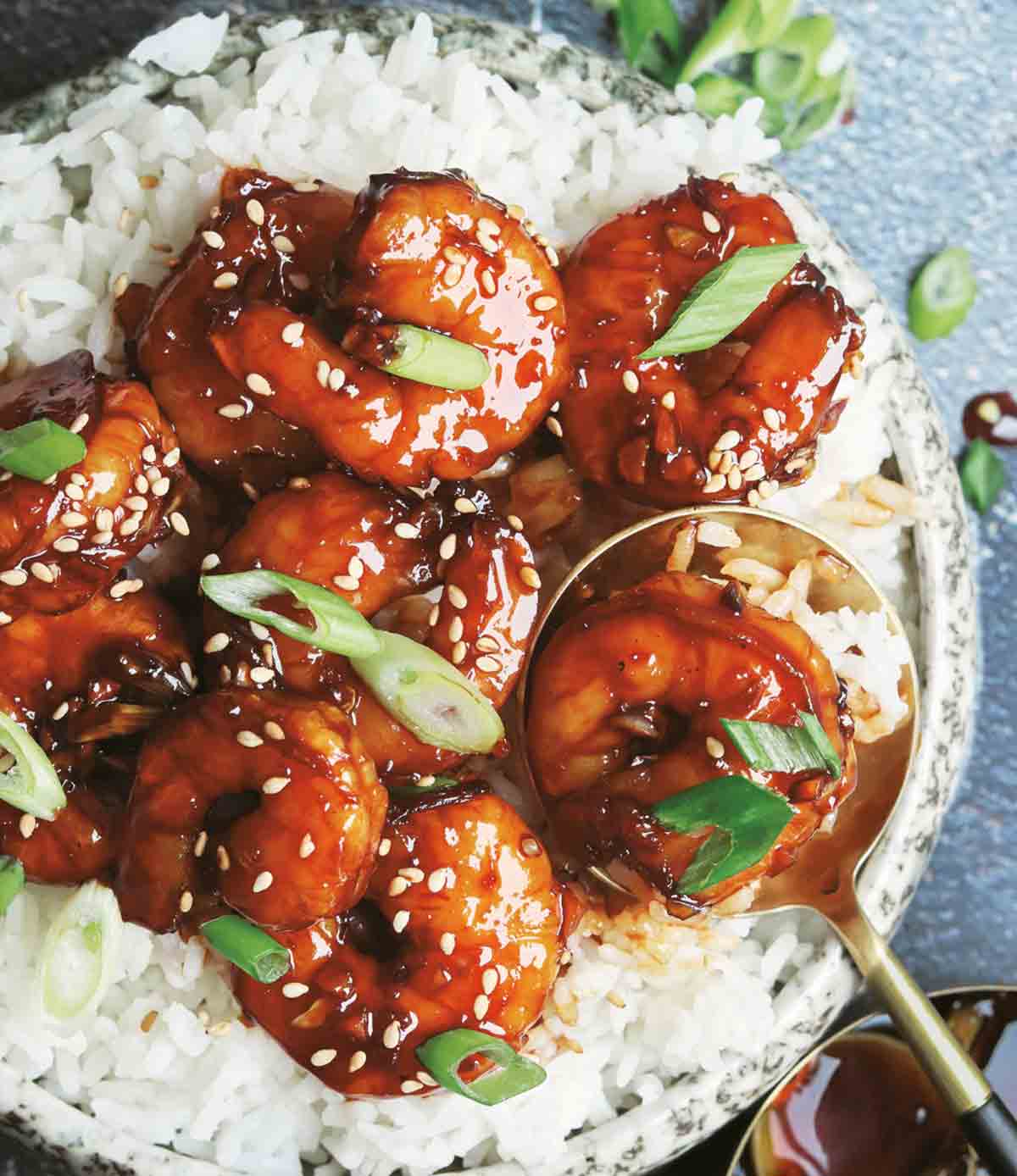 Glazed shrimp on top of rice and garnished with scallions, with a gold spoon scooping some from the bowl.