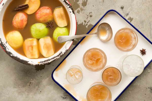 A chipped pot filled with apple pie moonshine, apple wedges and whole spices with a tray of glasses filled with the moonshine beside it.