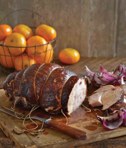 A brandy-brined turkey breast on a wooden cutting board with a couple slices cut off, a basket of mandarins in the background, and some grilled red onion wedges scattered around.