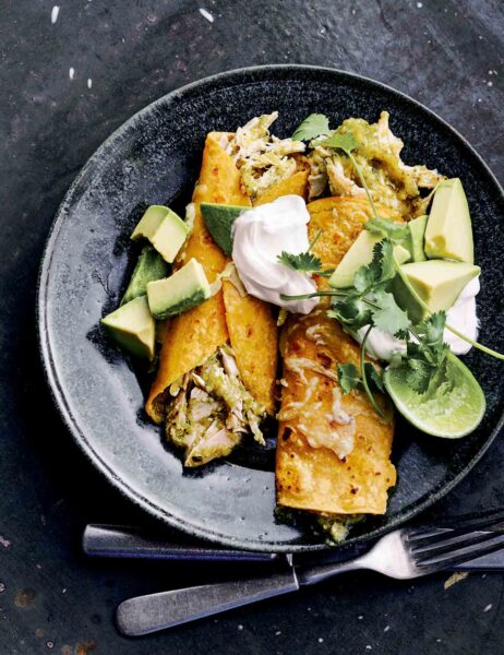 Two chicken enchiladas with green sauce in a black shallow bowl with cubed avocado, sour cream, cilantro, and a lime half.