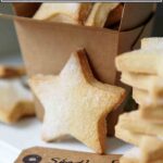 A stack of star-shaped cutout classic shortbread cookies, lightly dusted with sugar, and a brown takeout container and a gift tag.