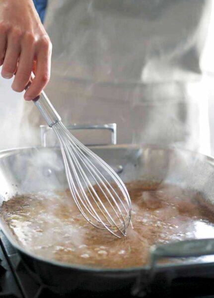 A person whisking gravy with a large metal balloon whisk in a metal roasting pan.