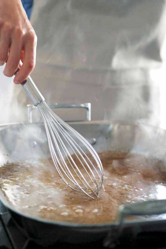 A person whisking gravy with a large metal balloon whisk in a metal roasting pan.