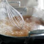 A person whisking gravy with a large metal balloon whisk in a metal roasting pan.