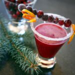 A cranberry margarita in a short glass with a skewer or sugared cranberries and orange zest across the top.