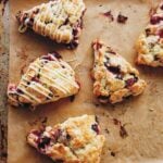 Five cranberry scones with two drizzled with white chocolate glaze on a parchment-lined rimmed baking sheet.