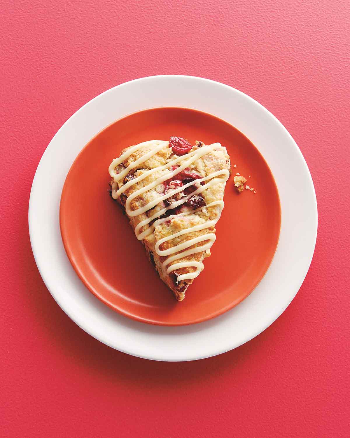 A cranberry scone with white chocolate drizzle on a red plate, which is set on a white plate on a red background.