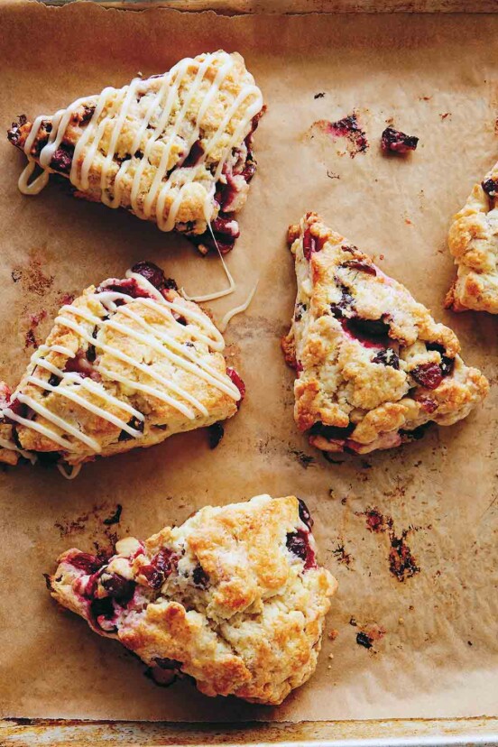 Five cranberry scones with two drizzled with white chocolate glaze on a parchment-lined rimmed baking sheet.
